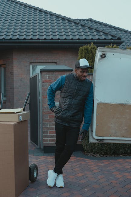 A young man wearing a beige knit beanie, a green puffer vest over a navy blue jacket with white stripes on the sleeves, and black gloves, sits on the edge of an open van in a residential driveway duri
