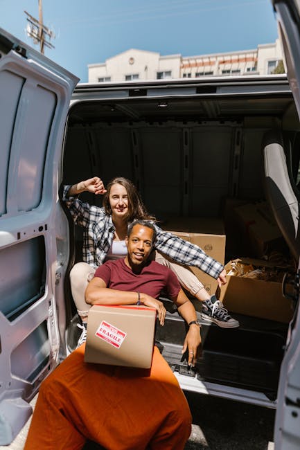 Two women smiling and posing inside the open rear cargo area of a van, which is loaded with cardboard moving boxes of various sizes, some sealed with packing tape and marked with fragile labels. One w